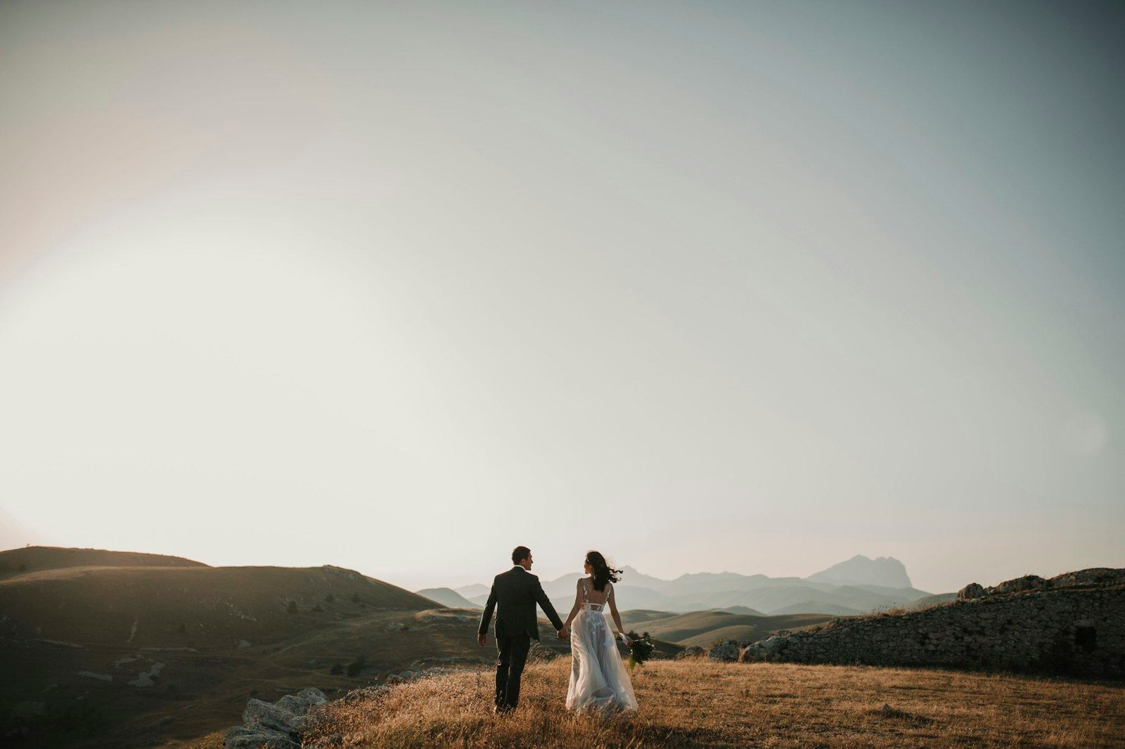 Pasangan pengantin bergandengan tangan berjalan di padang rumput luas, pegunungan dan langit berkabut di belakang mereka