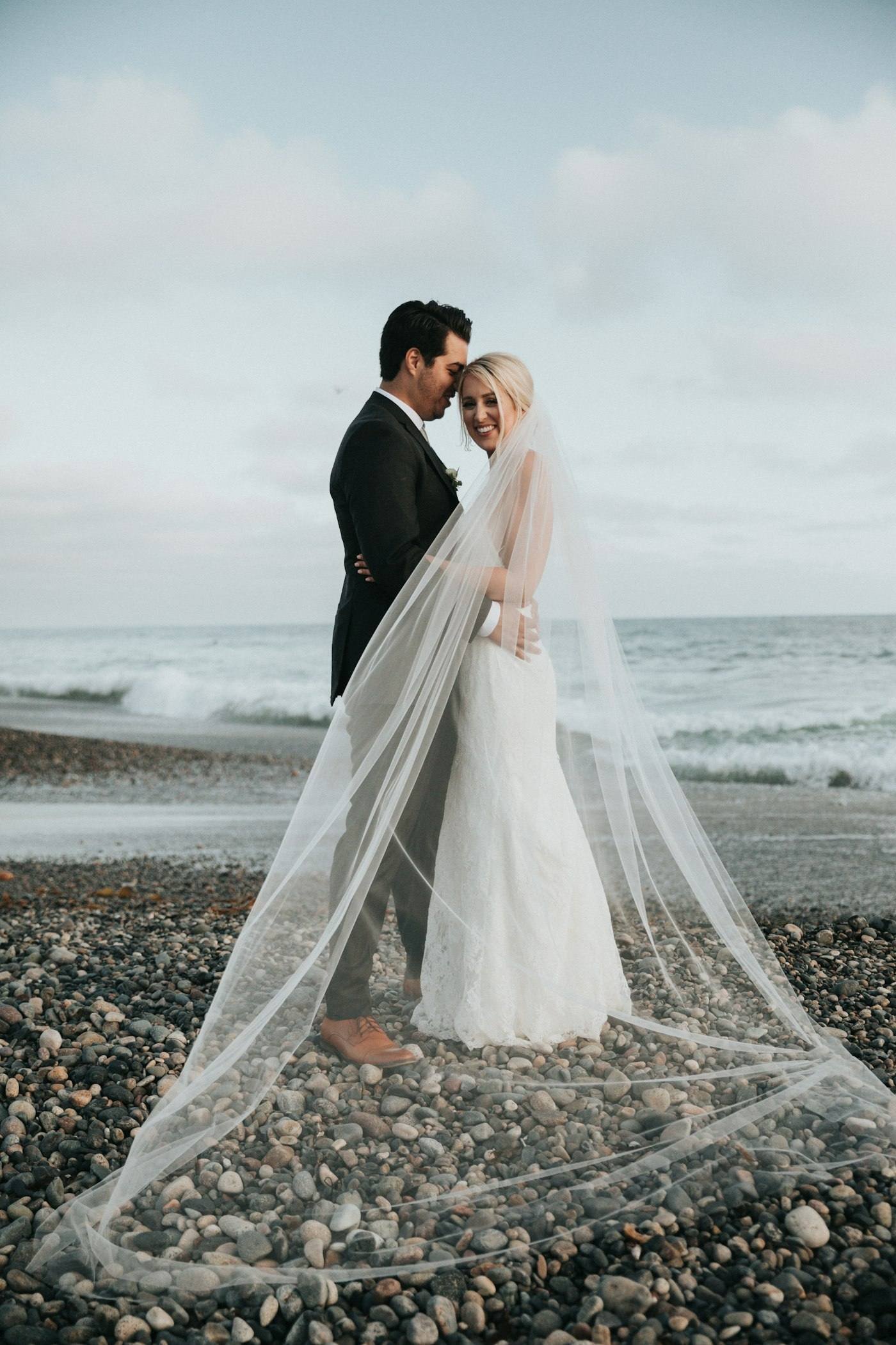 Pengantin berpelukan di pantai berbatu, kerudung panjang terbentang di atas kerikil